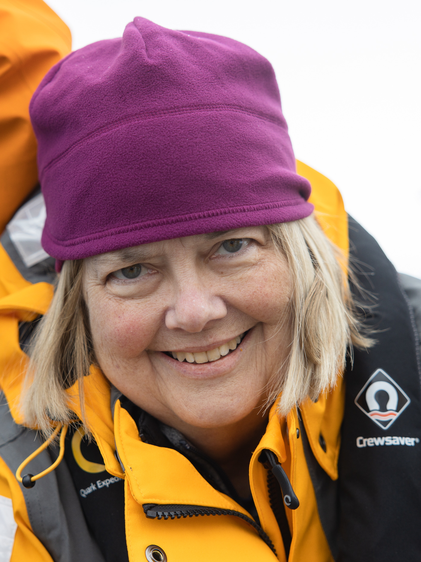 Photo of Margaret on January 14, 2023, standing on Antarctica. She has a yellow parka, a pink hat, and a big smile.